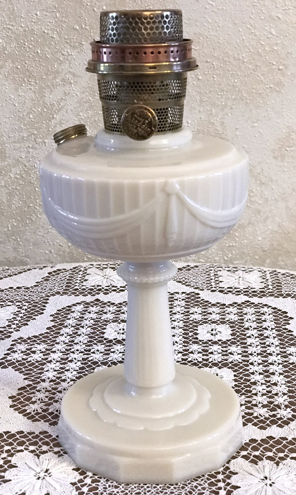 White vintage mantle oil lamp with ornate base and metal burner on a lace tablecloth.