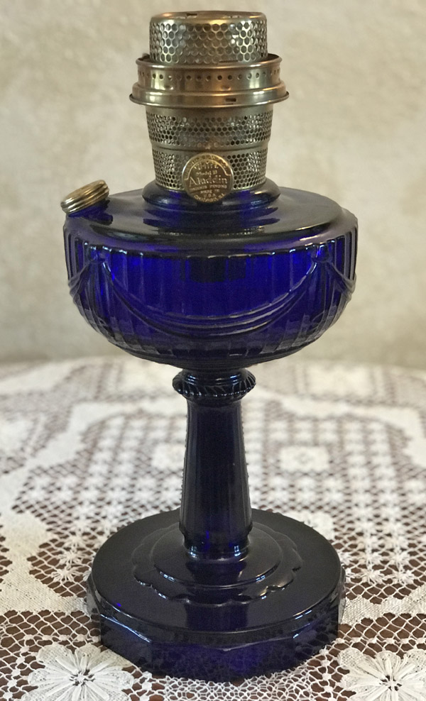 Tall cobalt blue glass oil lamp with scalloped design on a lace tablecloth.