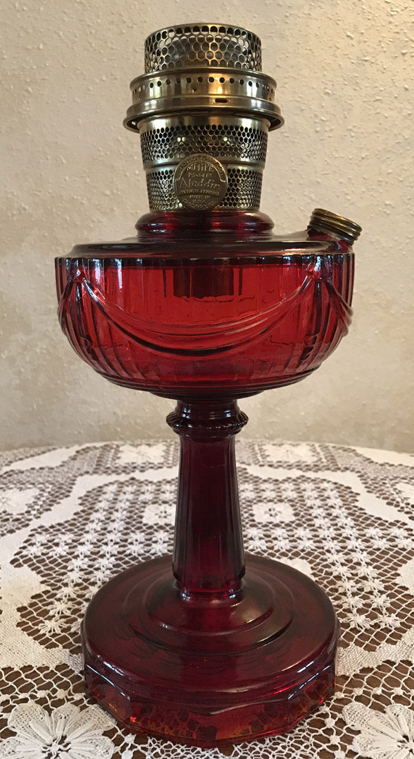 Tall vintage red glass oil lamp with brass burner on a lace tablecloth.