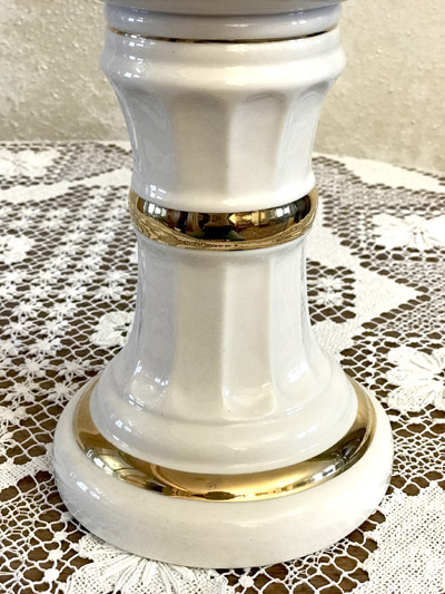 White ceramic pedestal with gold bands sitting on a lace-patterned tablecloth.