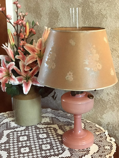 Pink vintage table lamp with floral shade on lace tablecloth next to pink lilies in a vase.