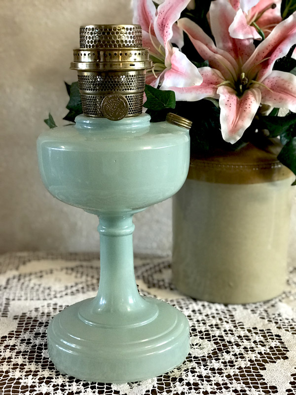 Light green vintage oil lamp with brass burner standing on lace tablecloth, pink lilies in background.