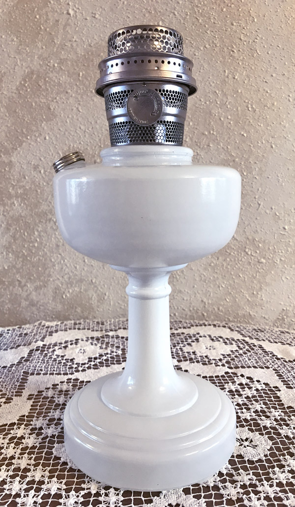White vintage oil lamp with metal burner sitting on lace tablecloth against textured wall.