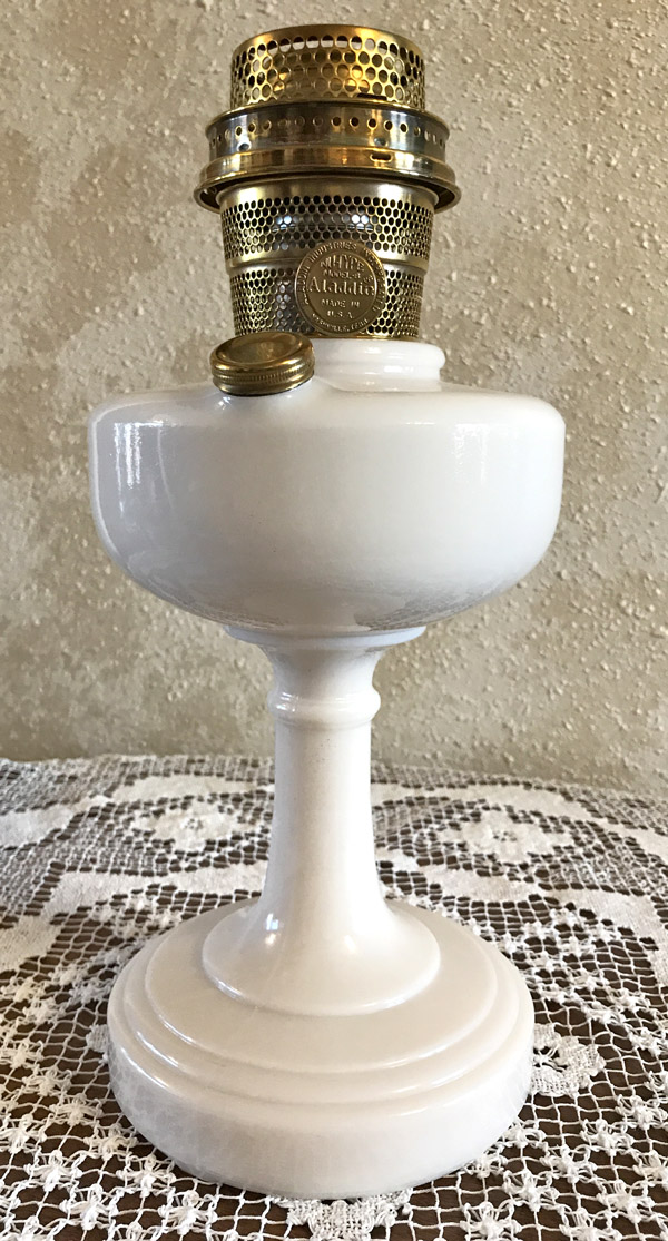White antique Aladdin oil lamp with brass burner on a lace tablecloth against a textured wall.