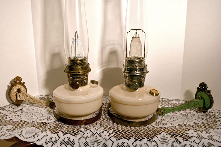 Pair of vintage kerosene lamps with cream bases on a lace tablecloth with decorative metal handles.