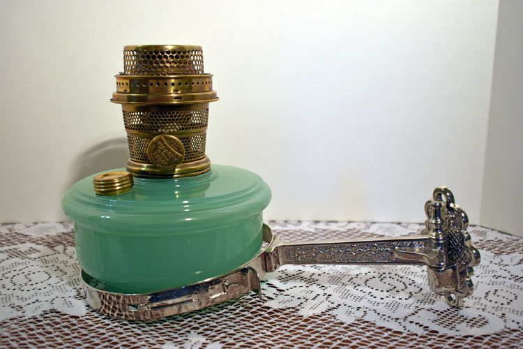 Antique green glass oil lamp with brass burner and ornate silver metal handle on lace tablecloth.