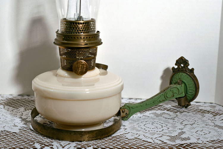 Antique oil lamp with white glass base and green decorative metal handle on lace-covered surface.