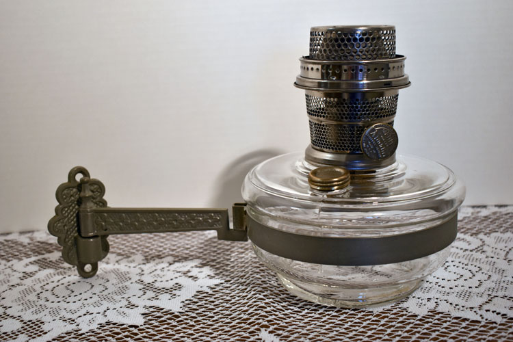 Vintage clear glass oil lamp with ornate metal burner and attached metal handle on lace surface.