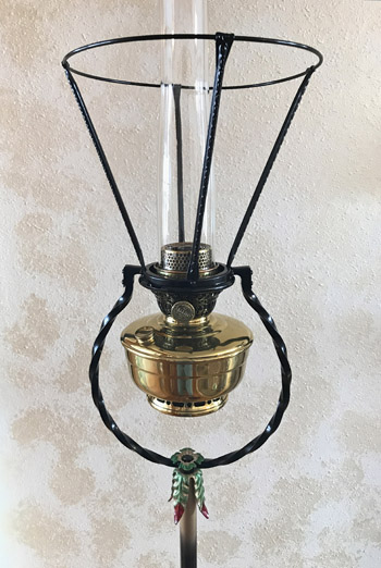 Antique brass oil lamp with glass chimney and twisted metal frame against a beige wall.
