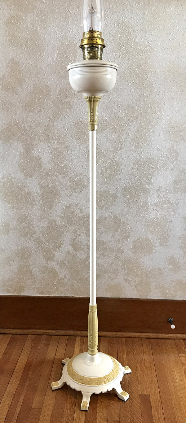 Tall vintage white and gold floor lamp with ornate base and glass chimney burner on hardwood floor.