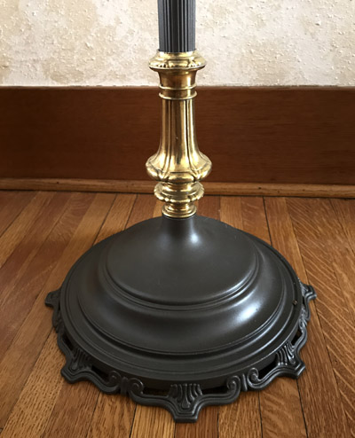 Base of a decorative lamp with a black ornate bottom and a gold midsection on a wood floor.