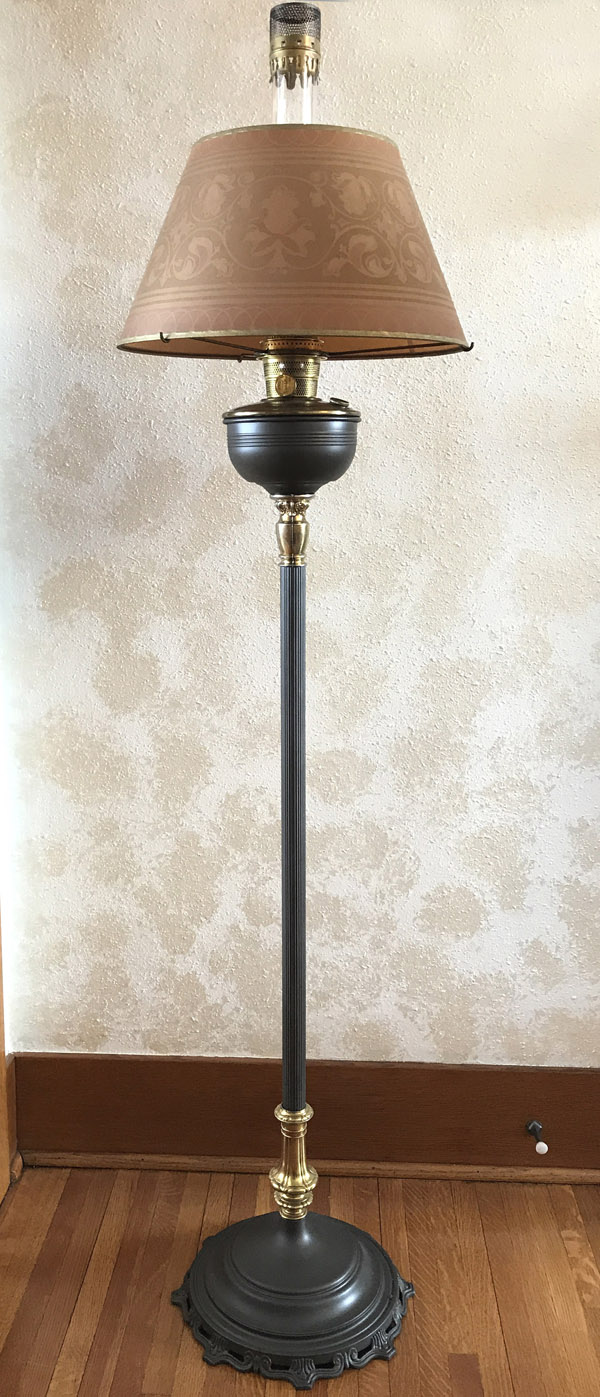 Tall vintage standing oil lamp with a brown patterned lampshade against a textured wall.