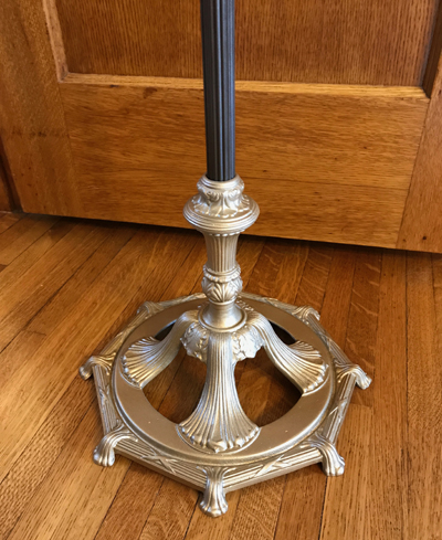 Ornate gold-colored lamp base with detailed leaf and fan patterns on a wood floor in front of a wooden door.