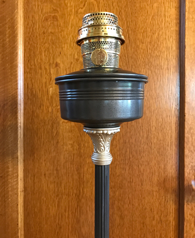 Close-up of a metal oil lamp burner on a dark stand against a wooden background.