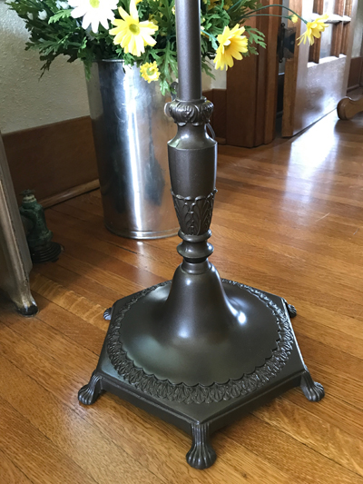Metal lamp base with decorative patterns standing on a wooden floor near a metal vase with yellow flowers.