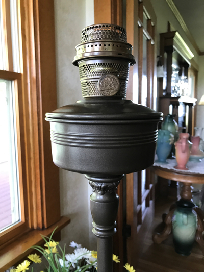Close-up of a vintage Aladdin oil lamp burner on a stand near a window with floral decor nearby.