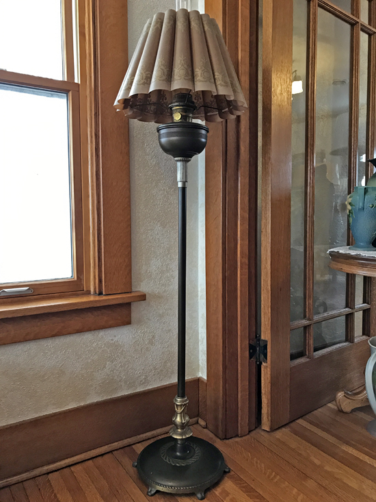Vintage floor lamp with a pleated beige lampshade standing on wooden floor by a window and door.