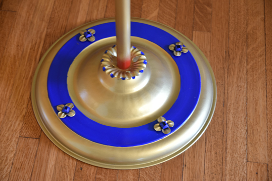 Metal lamp base with blue ring and decorative floral accents on a wooden floor.