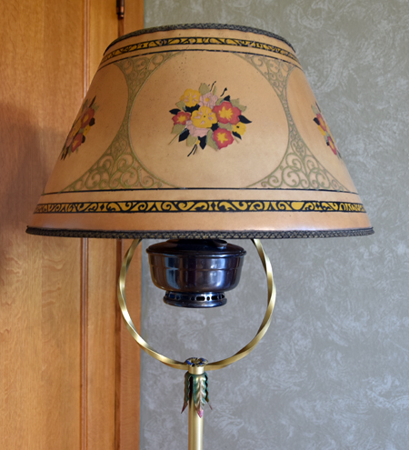 Antique lamp with floral-decorated beige shade and brass ring stand against textured and wooden wall.