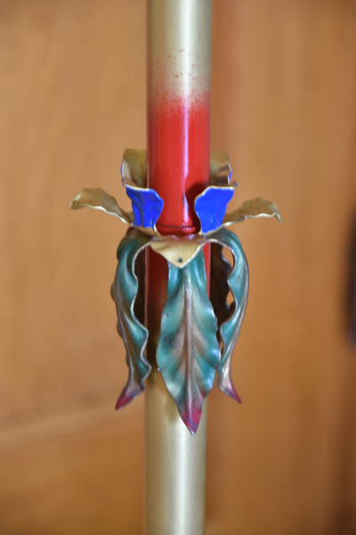 Red candle with decorative metal holder shaped like leaves and blue petals on a gold stand.