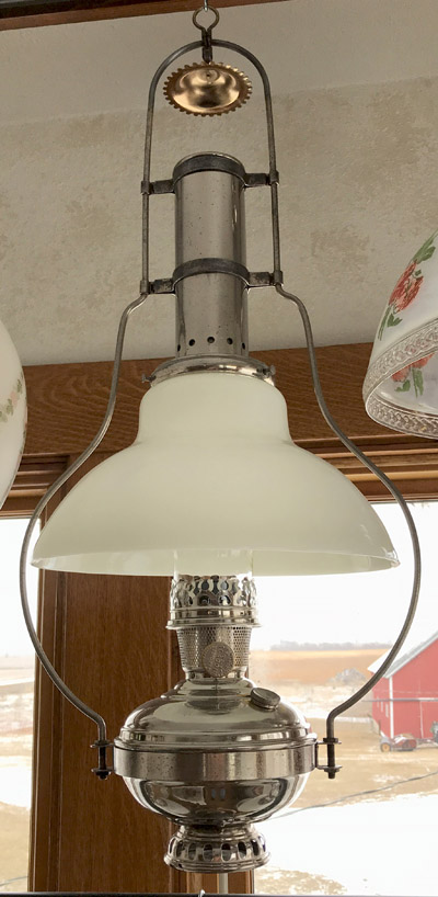 Vintage metal and glass hanging oil lamp with a white shade by a window showing a snowy farm scene.