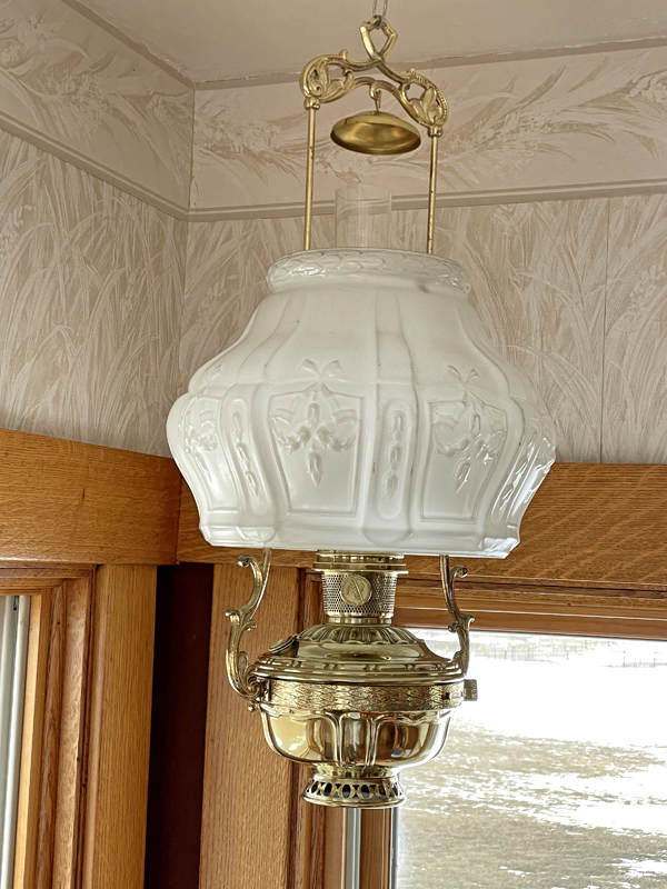 Vintage brass and white embossed glass hanging oil lamp near wooden window and wallpapered wall.