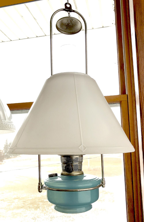 Vintage blue oil lamp with a white conical shade hanging by a window with a wooden frame.