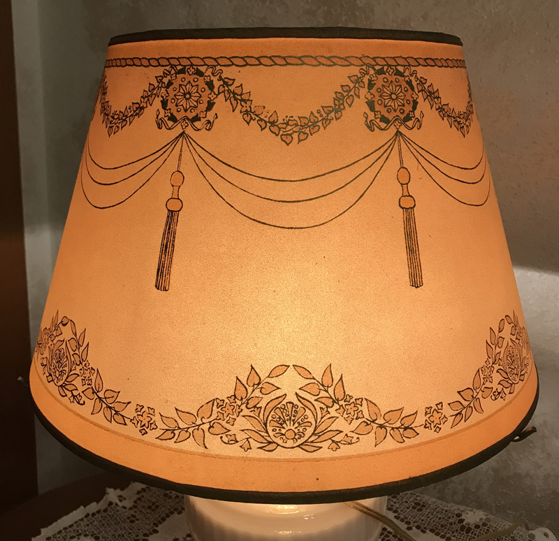 Illuminated lampshade with floral and tassel patterns in brown on warm orange background.