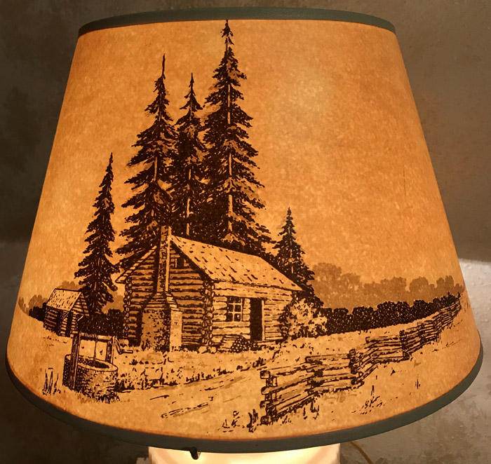 Lamp shade with an illustration of a cabin, tall pine trees, fence, and a water well.