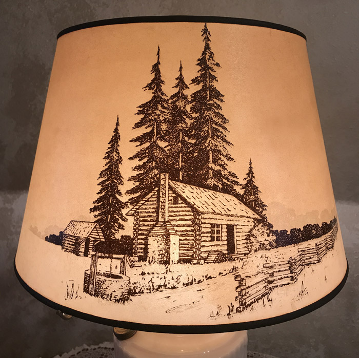 Lamp shade with rustic scene of log cabin, tall pine trees, wooden fence, and a well.