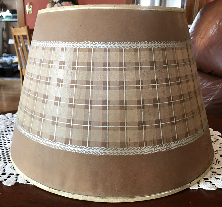 Brown lampshade with beige plaid pattern and white leaf borders sitting on a wooden table with a lace cloth.