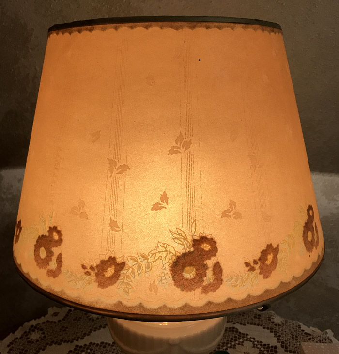 Lit beige lampshade with brown floral patterns and scalloped edges on a white ceramic lamp base.