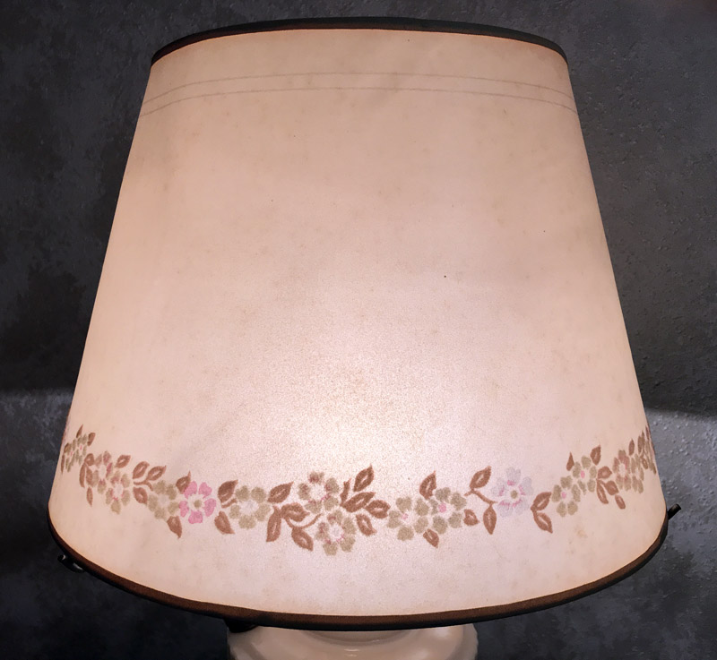 Beige lamp shade with a floral pattern border near its bottom edge against a dark background.