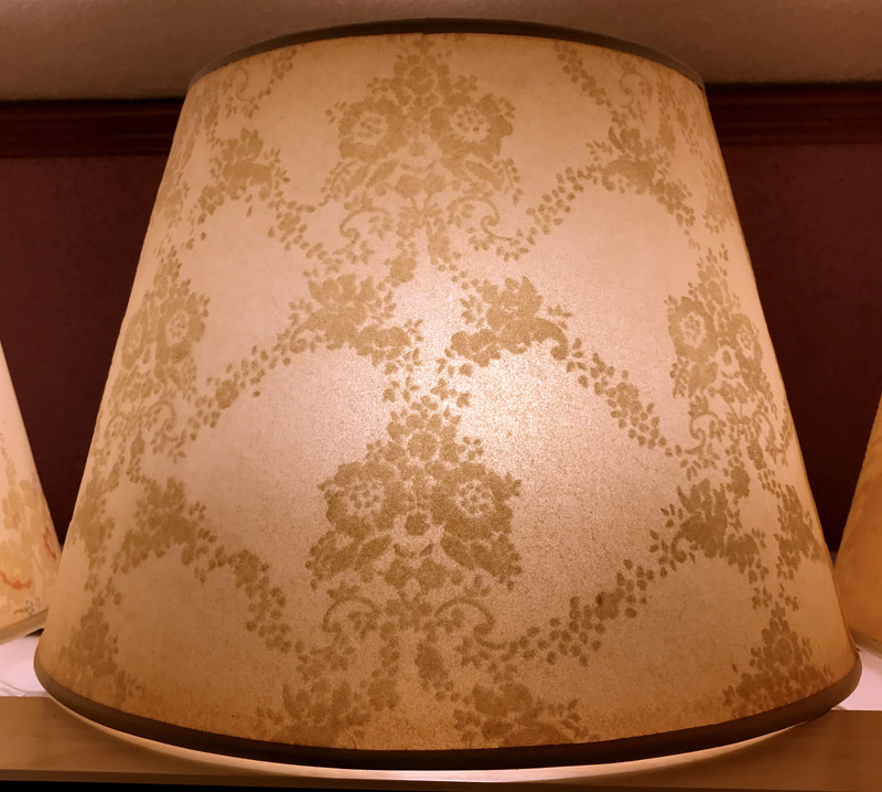 Beige lampshade with a subtle floral damask pattern and brown trim edges sitting on a wooden surface.