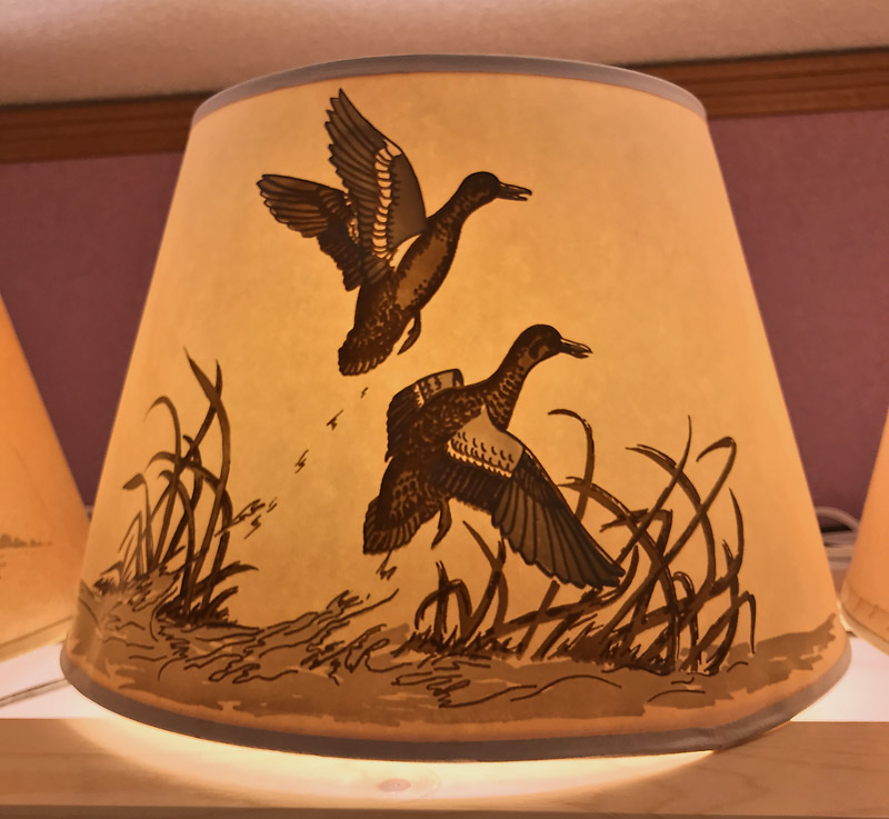 Lampshade with illustration of two flying ducks over marsh grasses, glowing warmly.