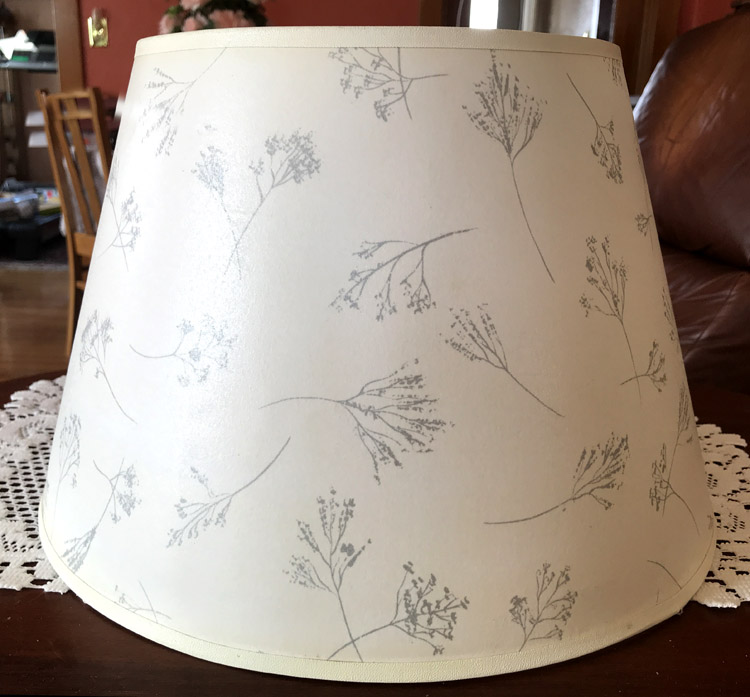 Off-white lampshade with delicate gray botanical sketches on a wooden surface with lace doily.