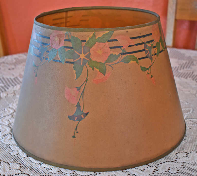Brown lampshade with green and pink morning glory flowers painted on a music staff, on a lace tablecloth.