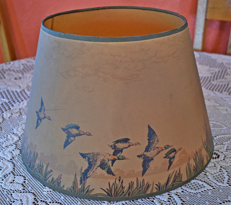 Beige lampshade with painted flying ducks and tall grasses sitting on a lace tablecloth.