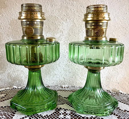 Two vintage green glass oil lamps with brass burners on a lace tablecloth.