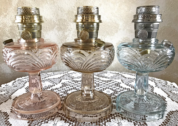 Three vintage glass oil lamps in pink, clear, and blue on a lace tablecloth against a beige wall.