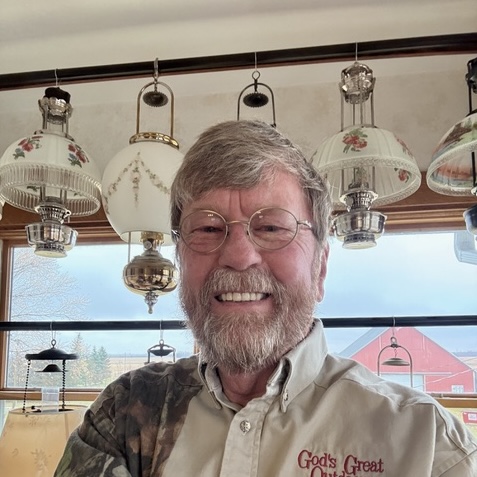 Smiling bearded man wearing glasses and a tan shirt embroidered with 'God's Great Light'.