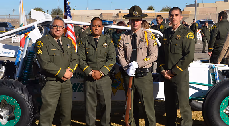 Four uniformed law enforcement officers standing in front of a vehicle at an outdoor event.