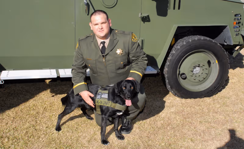 Uniformed officer kneeling beside a black K-9 unit dog in front of a green military vehicle.