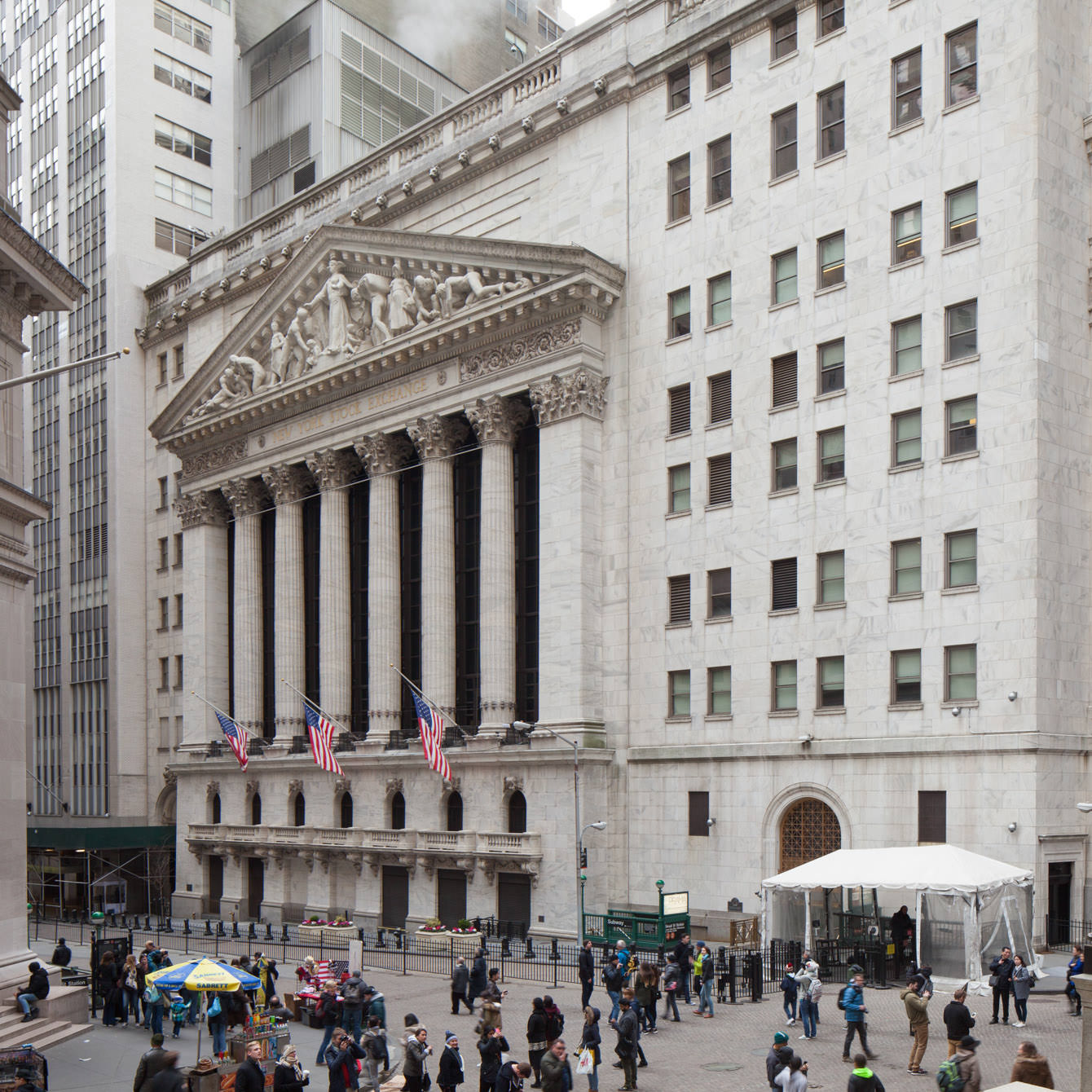 People walking on Wall Street with tall buildings and American flags on the New York Stock Exchange.
