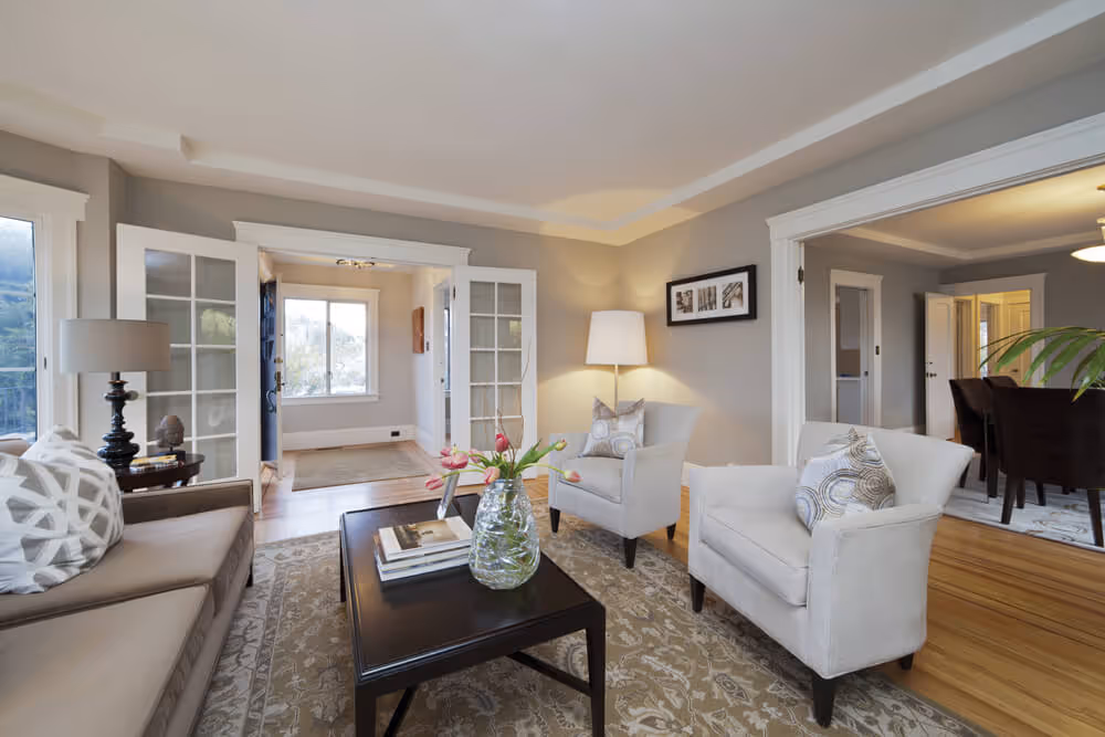 Bright living room with beige sofa, two cushioned armchairs, a dark wooden coffee table with a glass vase of pink tulips, and patterned area rug.