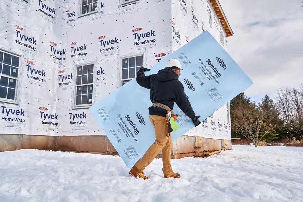 Construction worker carrying a blue DuPont Styrofoam insulation board outside a house wrapped in Tyvek HomeWrap during snowy weather.