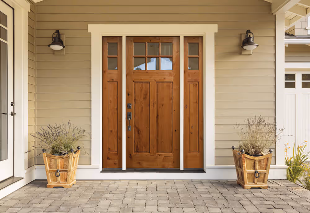 Wooden front door with glass panels framed by beige siding, flanked by two wooden planters with plants on a brick patio.