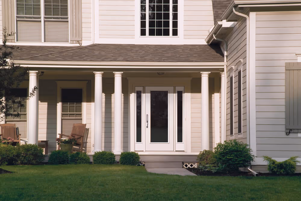 Front porch of a house with white columns, a white door with glass panels, and two wooden chairs surrounded by green bushes.