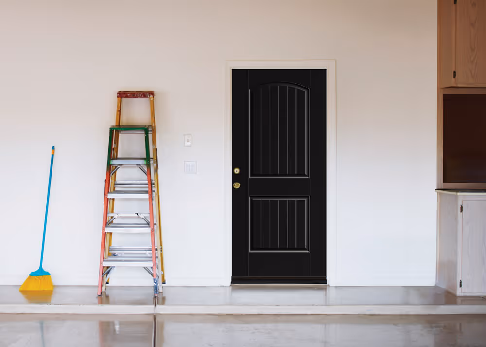 Black fire door with white frame in a plain garage wall next to a yellow broom and colorful stacked ladders.