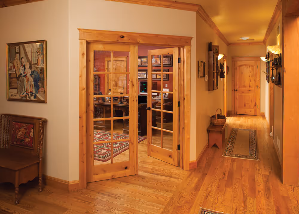 Hallway with wooden floors, featuring open wooden French doors leading to a cozy office with a patterned rug and wooden furniture.
